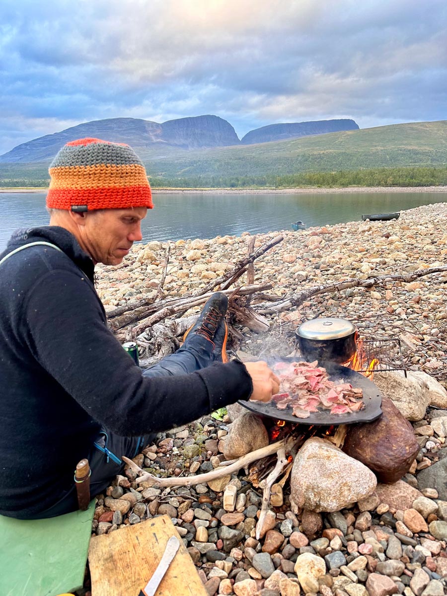 Person sitting by a small outdoor fire cooking on a rocky shoreline.