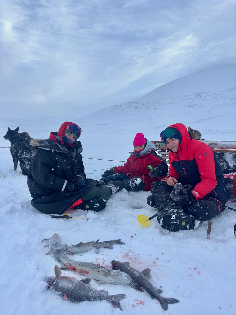 Three people dressed in winter clothing sitting on snowy ground with several fish lying on the snow in front of them, with a sled and two huskies in the background under a cloudy sky in a snowy mountainous landscape.