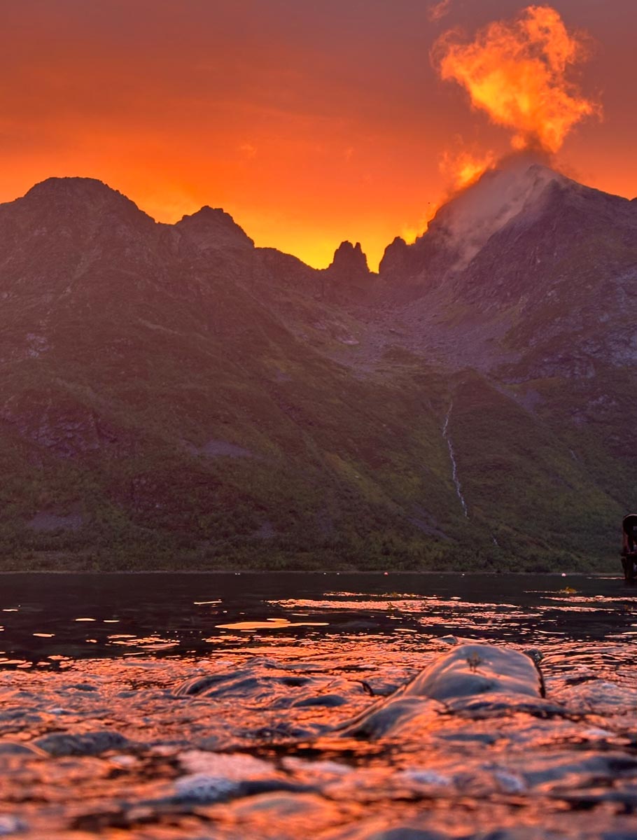 Fiery orange sunset casting a deep glow over sharp mountain peaks and water.