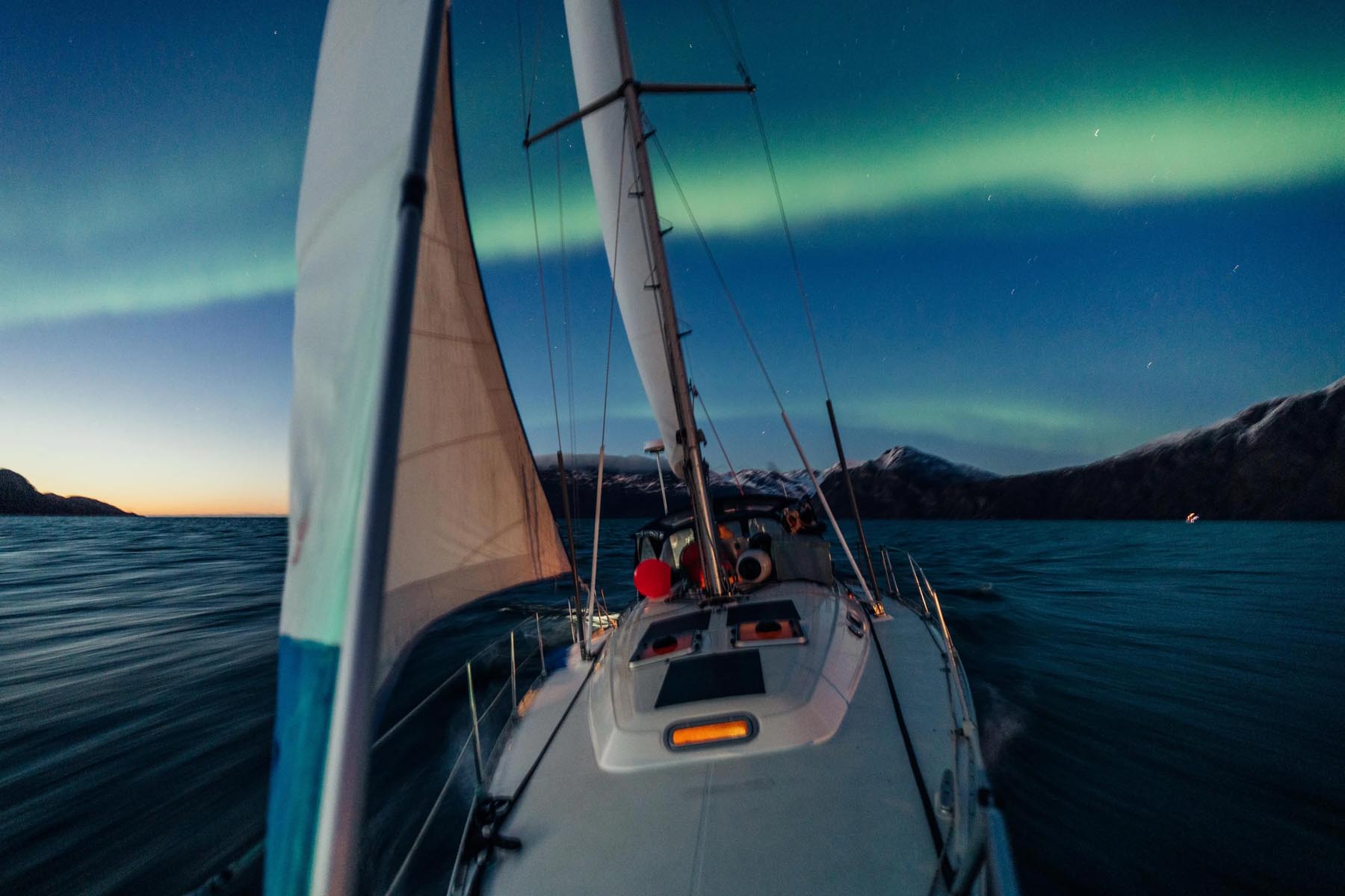 Sailboat bow heading forward beneath vibrant green northern lights at night.