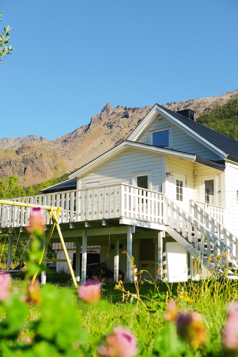 White wooden elevated house with a large porch and stairs, set against rocky mountains under a clear blue sky, with green grass and pink flowers in the foreground.