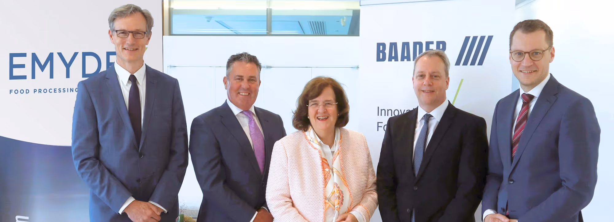 Five business professionals standing in front of banners with the logos EMYDEX and BAADER, all dressed in formal attire and smiling.