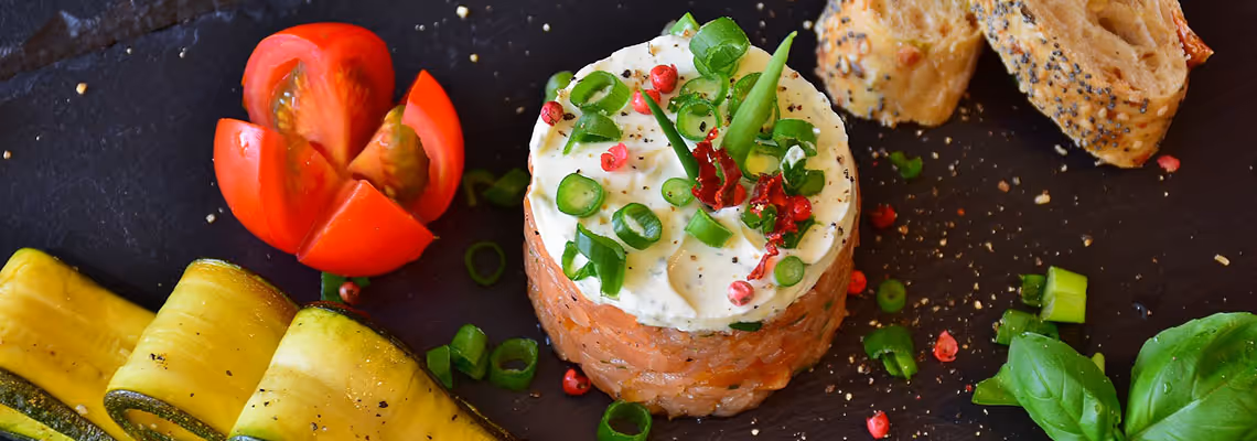 Salmon tartare topped with cream and chopped green onions, accompanied by sliced cherry tomato, rolled zucchini ribbons, toasted seeded bread, and basil leaves on a dark slate surface.