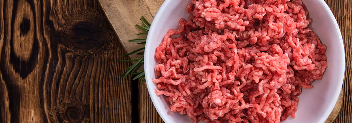 Close-up of minced raw meat in a white bowl on a wooden surface with a sprig of rosemary.