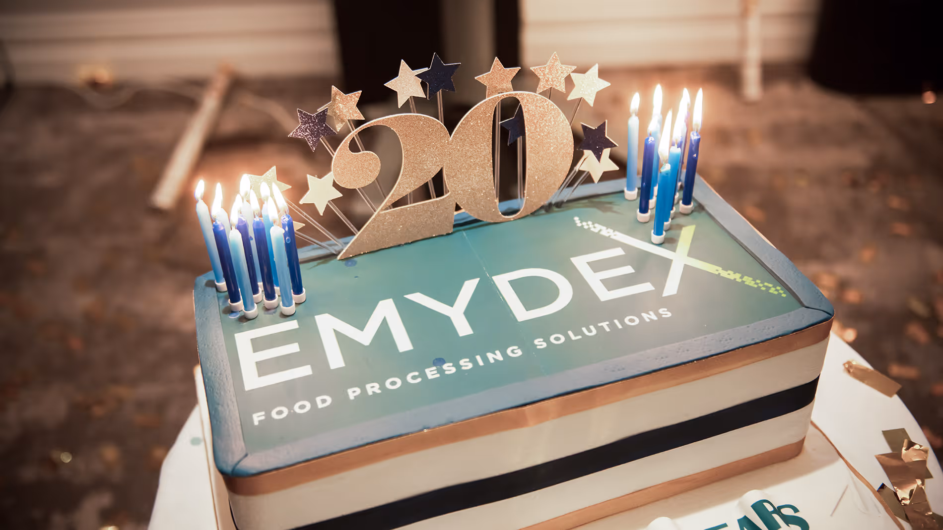 Rectangular celebration cake with EMYDEX Food Processing Solutions logo, topped with blue candles and a glittery '20' decoration with stars.