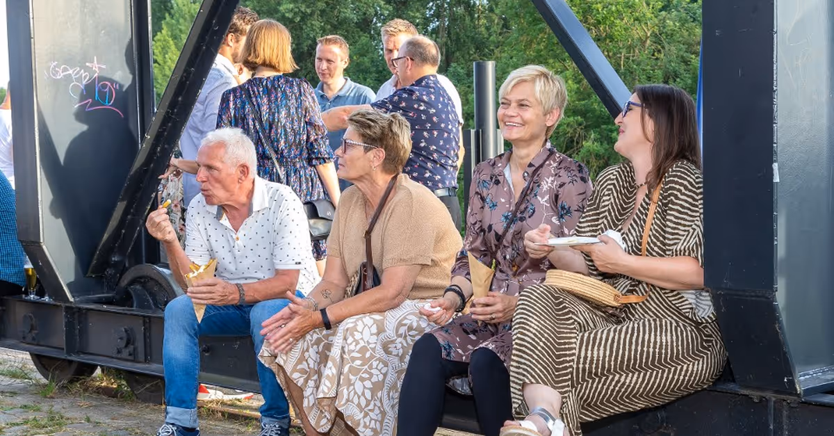 Four adults sitting on a metal beam outdoors eating snacks and chatting, with green trees in the background.