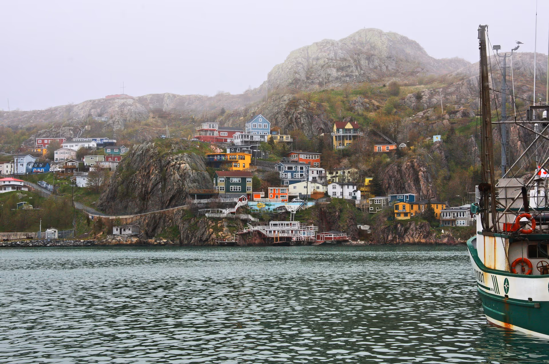 Colorful houses built on rocky hills overlooking calm water with a fishing boat partially visible on the right.