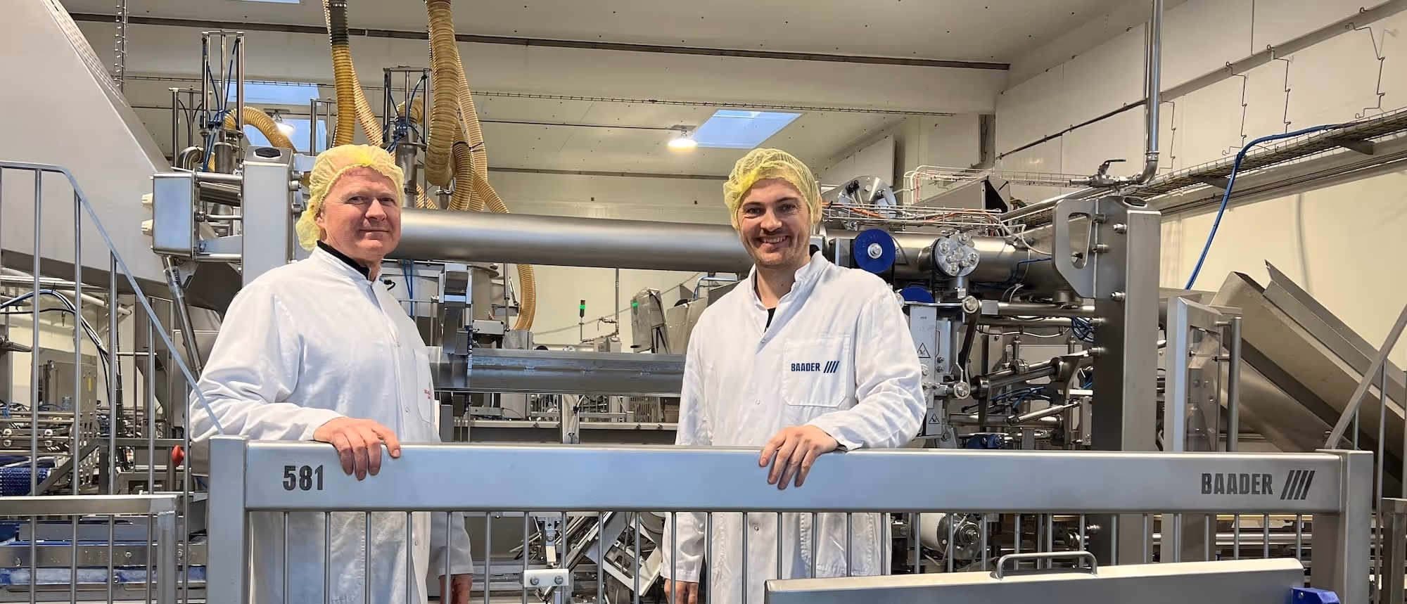 Two men in white lab coats and yellow hairnets standing behind metal railing in a food processing facility with industrial machinery.