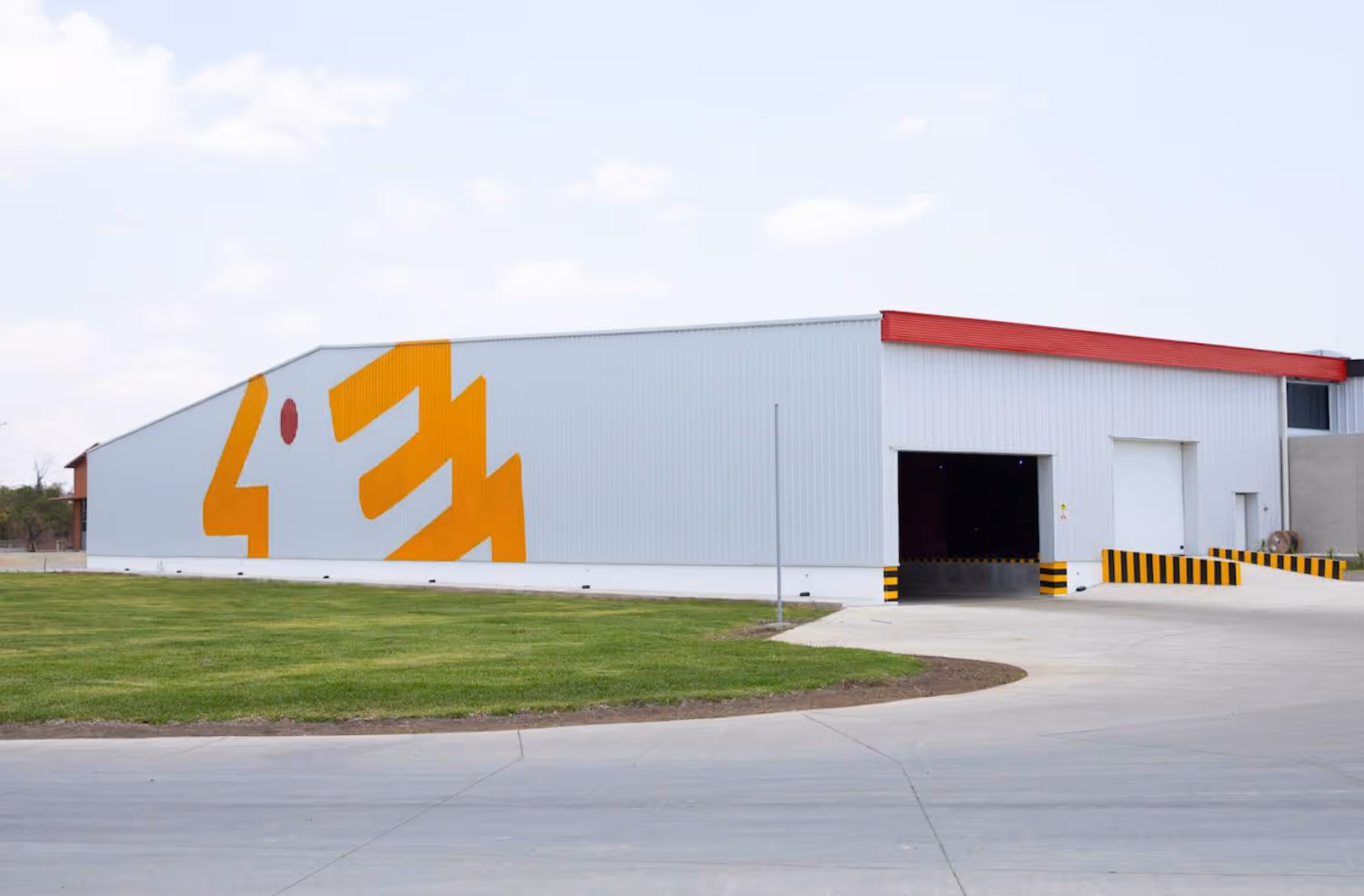 Large industrial warehouse with white and gray metal siding, a red roof edge, a yellow and orange geometric mural on the side, and open garage doors with yellow and black striped safety markings.
