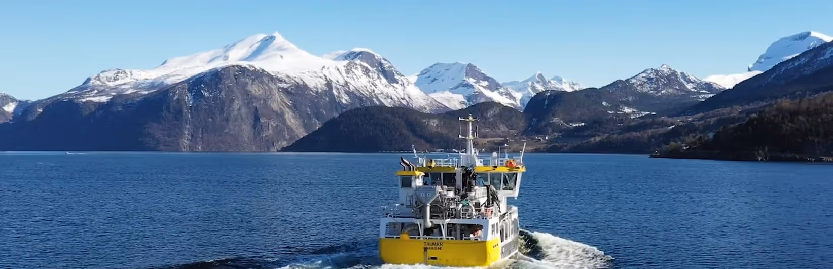 Yellow and white boat sailing on blue fjord waters with snow-capped mountains under a clear blue sky.
