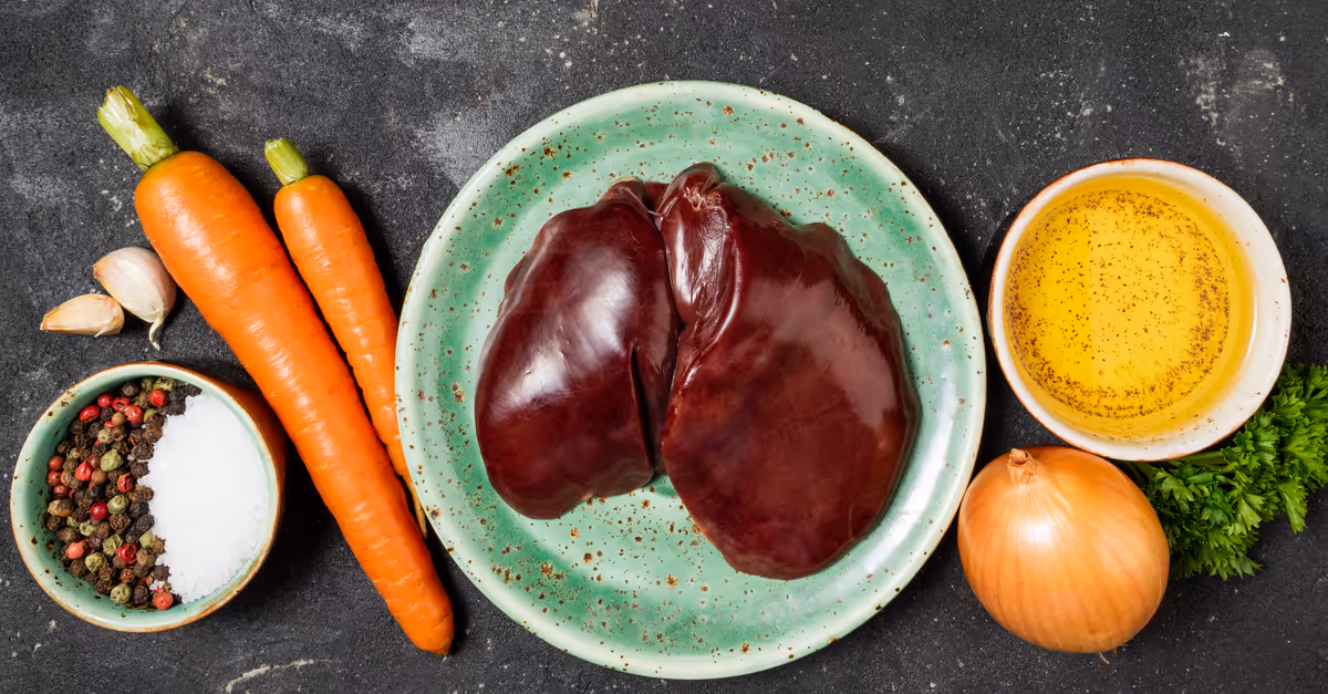 Raw liver on a green plate surrounded by carrots, garlic, onion, parsley, peppercorns, salt, and a bowl of oil with seasoning.