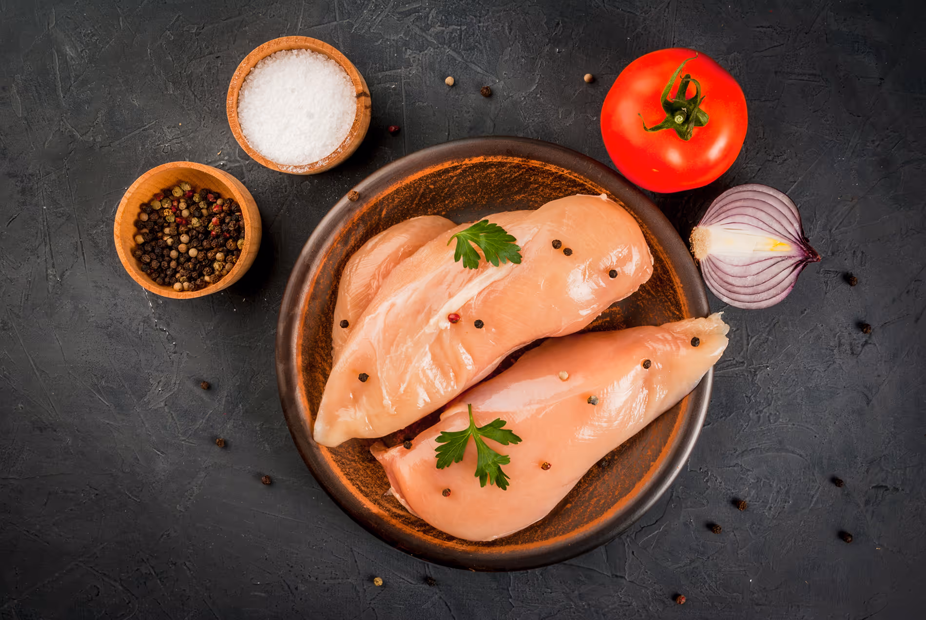 Raw chicken breasts on a brown plate garnished with parsley and peppercorns, surrounded by bowls of salt, mixed peppercorns, a tomato, and a halved red onion on a dark textured surface.