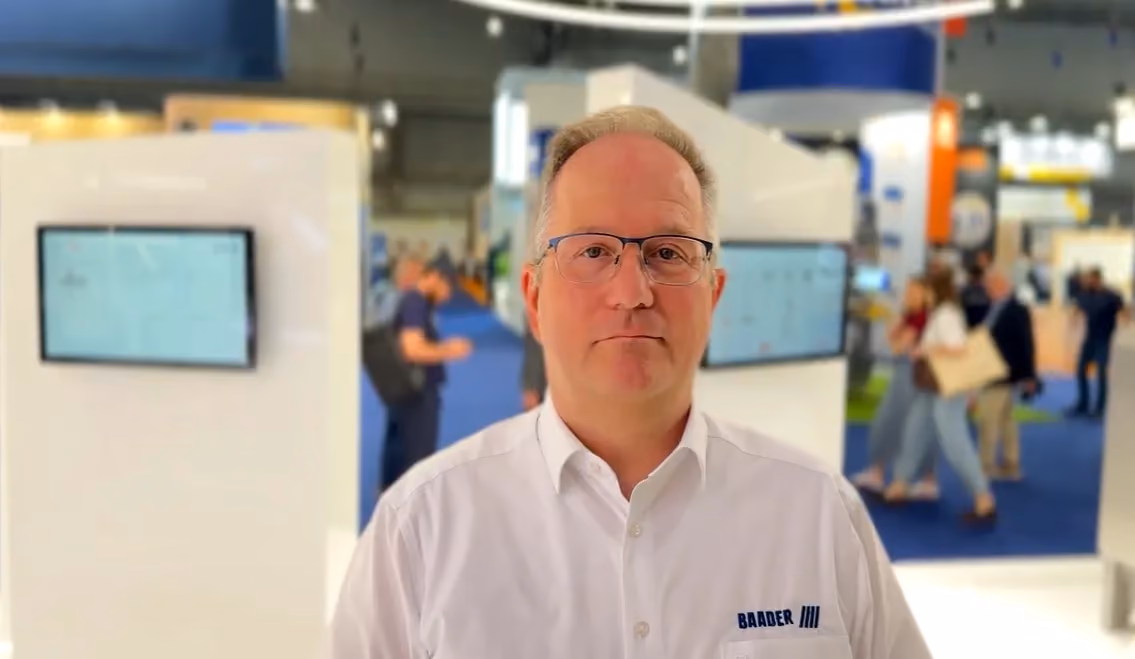 Middle-aged man wearing glasses and a white shirt with BAADER logo standing in a busy exhibition or trade show setting.