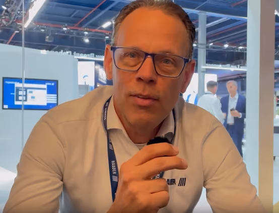 Man with glasses wearing a white shirt and lanyard speaking into a microphone indoors at a tech event.