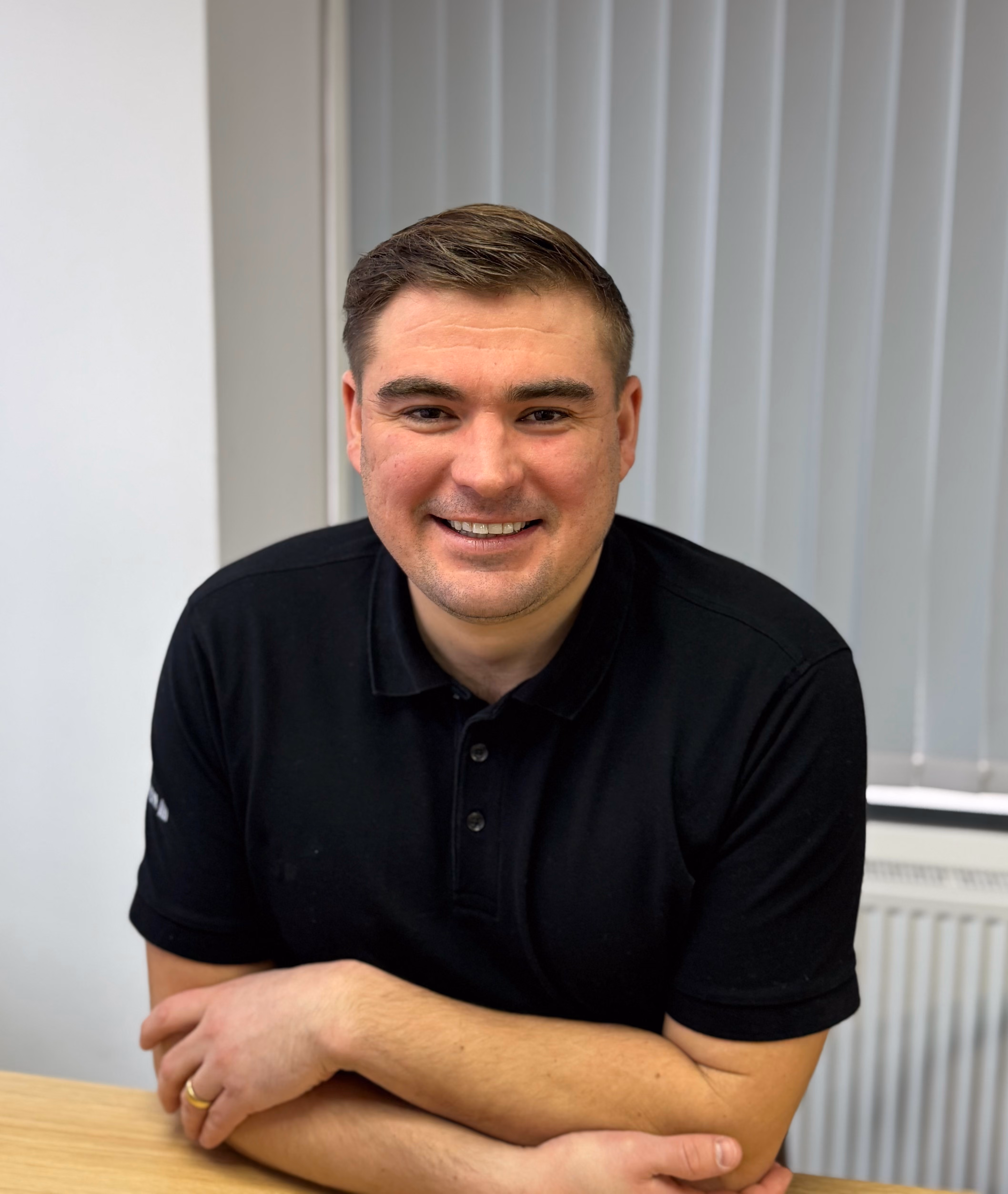 Smiling man in a black polo shirt sitting at a table with arms crossed.