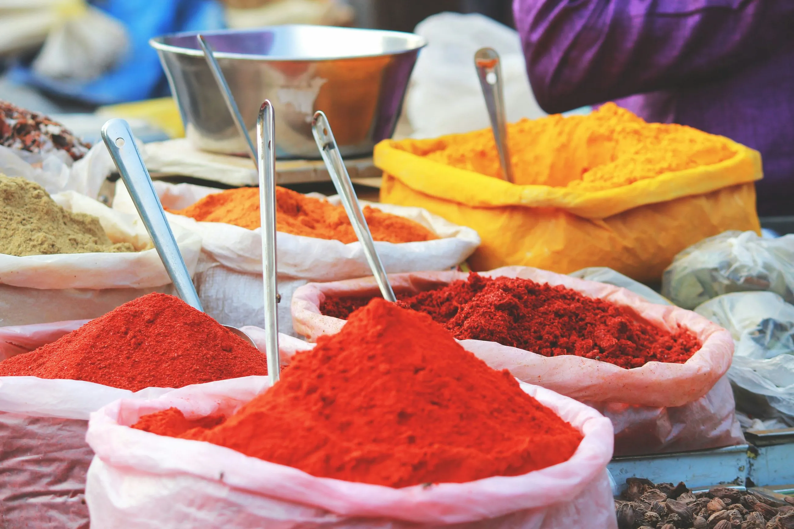Piles of colorful powdered spices including red, orange, and yellow hues displayed in open sacks with metal scoops in a market setting.