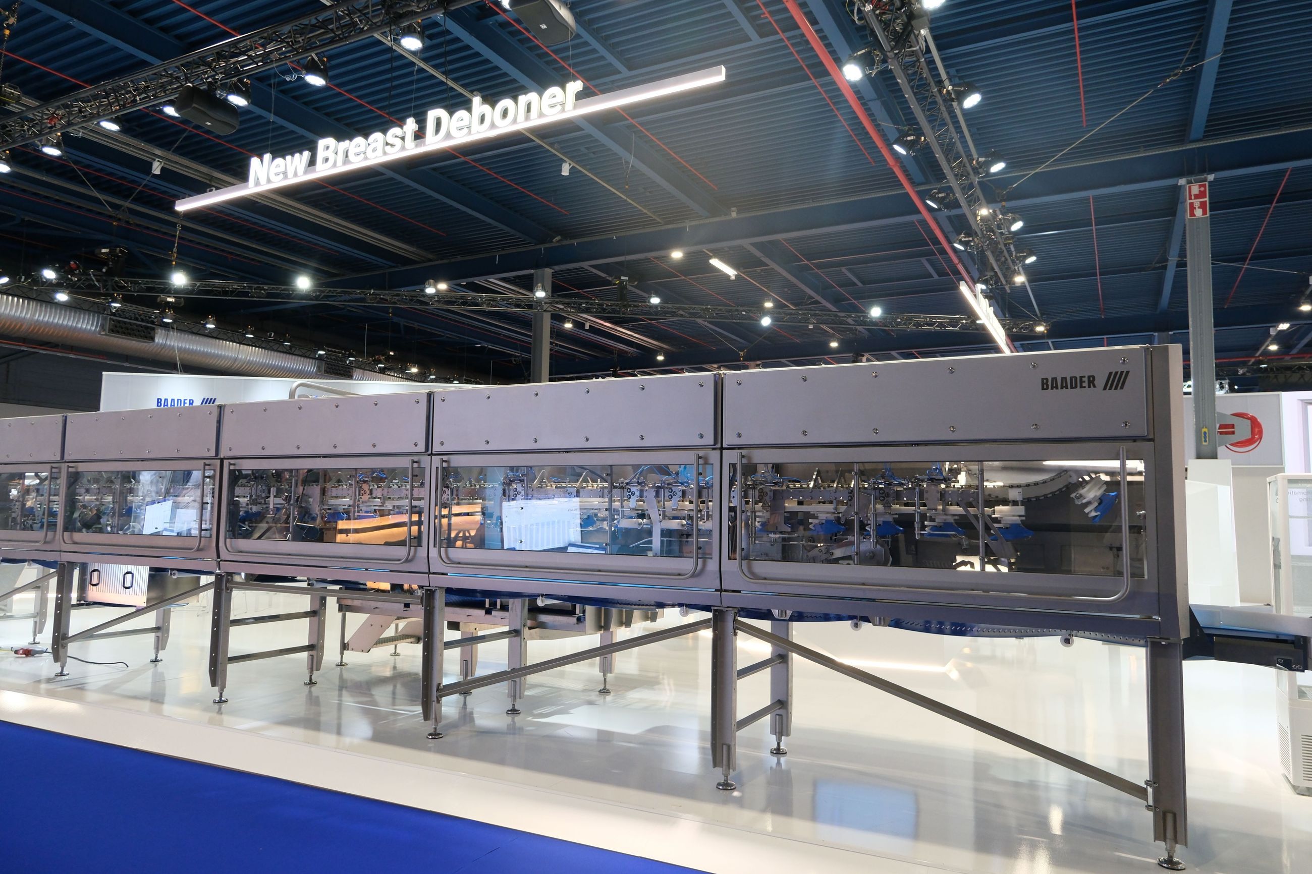 A large, industrial stainless steel meat processing machine labeled 'BAADER' on display in a well-lit exhibition hall with a sign above reading 'New Breast Deboner'.