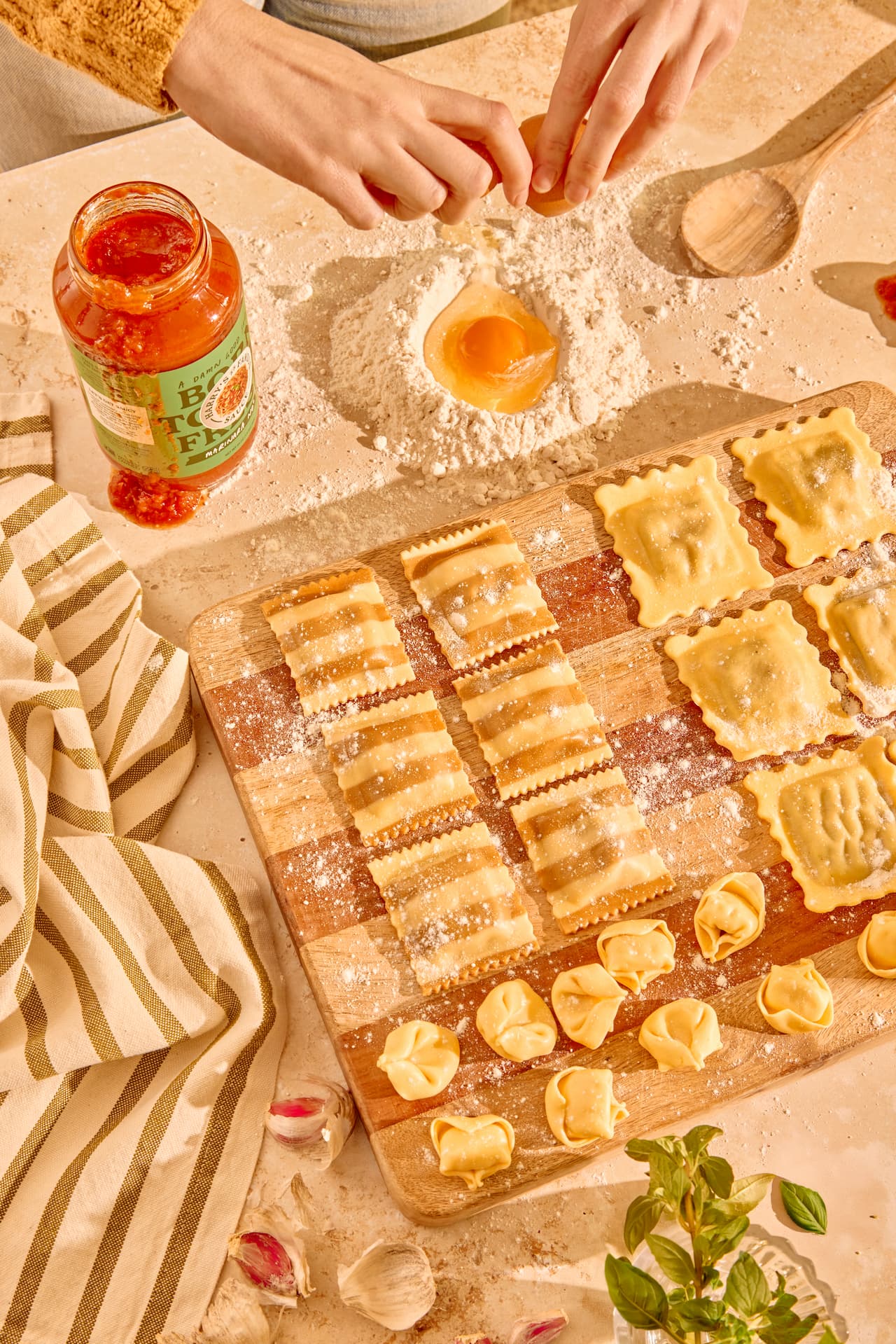 Hands cracking an egg into flour on a kitchen counter with a cutting board holding various shapes of homemade pasta, a jar of marinara sauce, garlic cloves, and a striped kitchen towel.