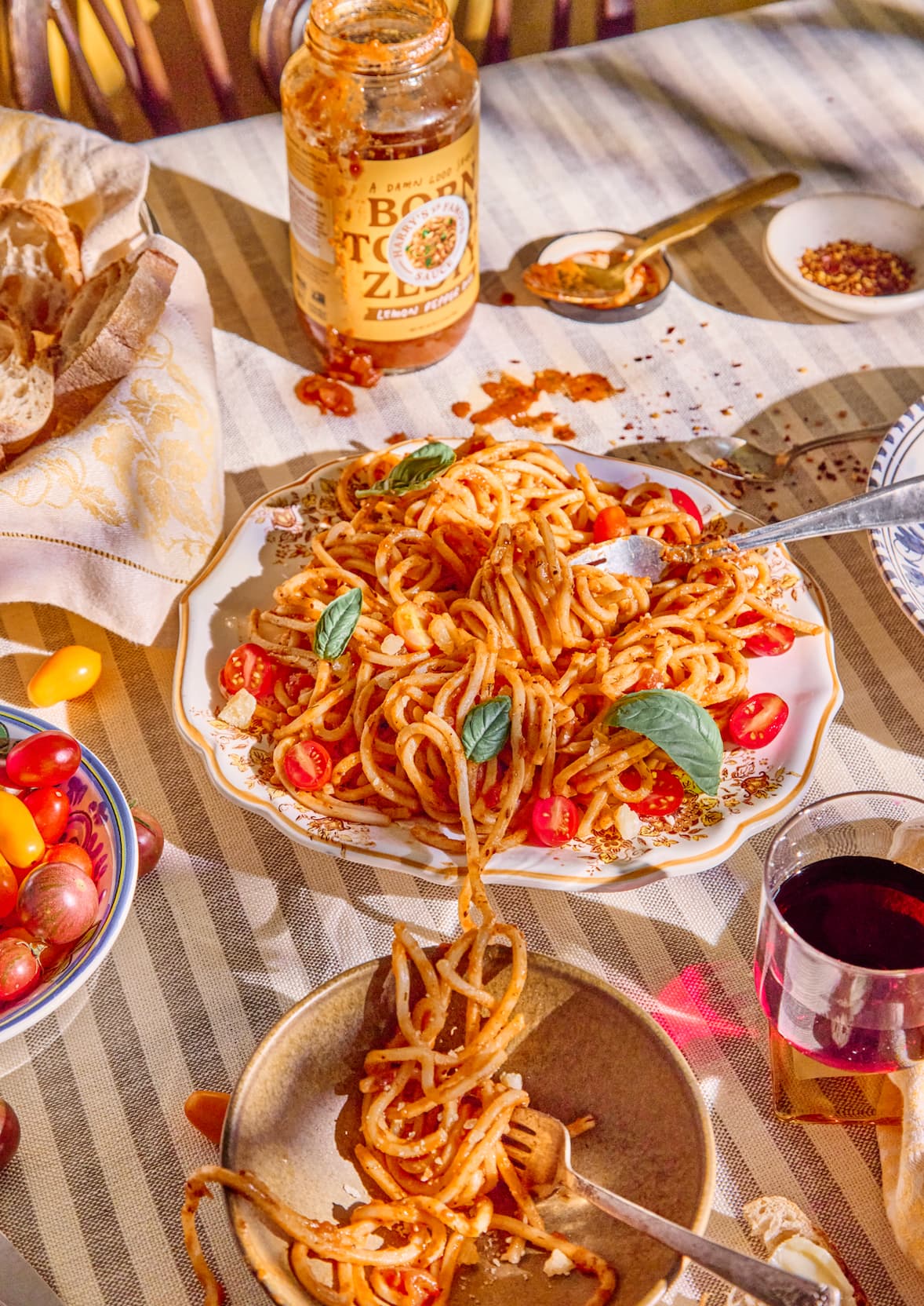 Plate of spaghetti with tomato sauce, cherry tomatoes, and basil on a striped tablecloth, with bread, a jar of sauce, and a glass of red wine nearby.