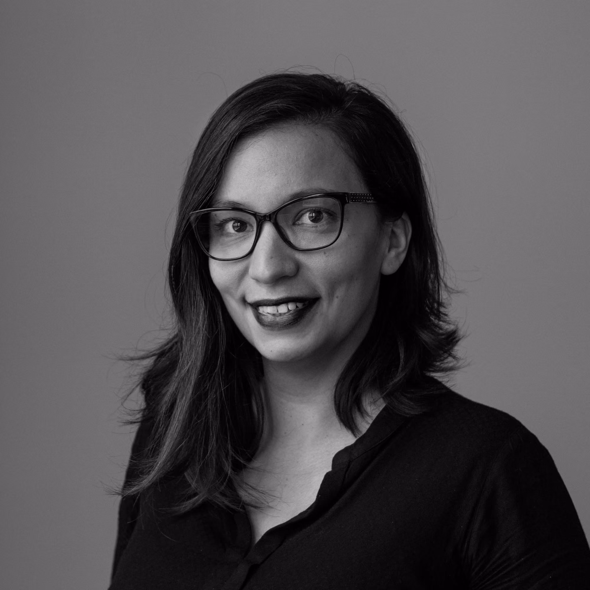 Smiling woman with glasses, medium-length hair, wearing a dark top against a plain background.