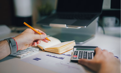Une personne prenant des notes dans un carnet avec un crayon, utilisant une calculatrice, avec un clavier et un ordinateur portable en arrière-plan sur un bureau.