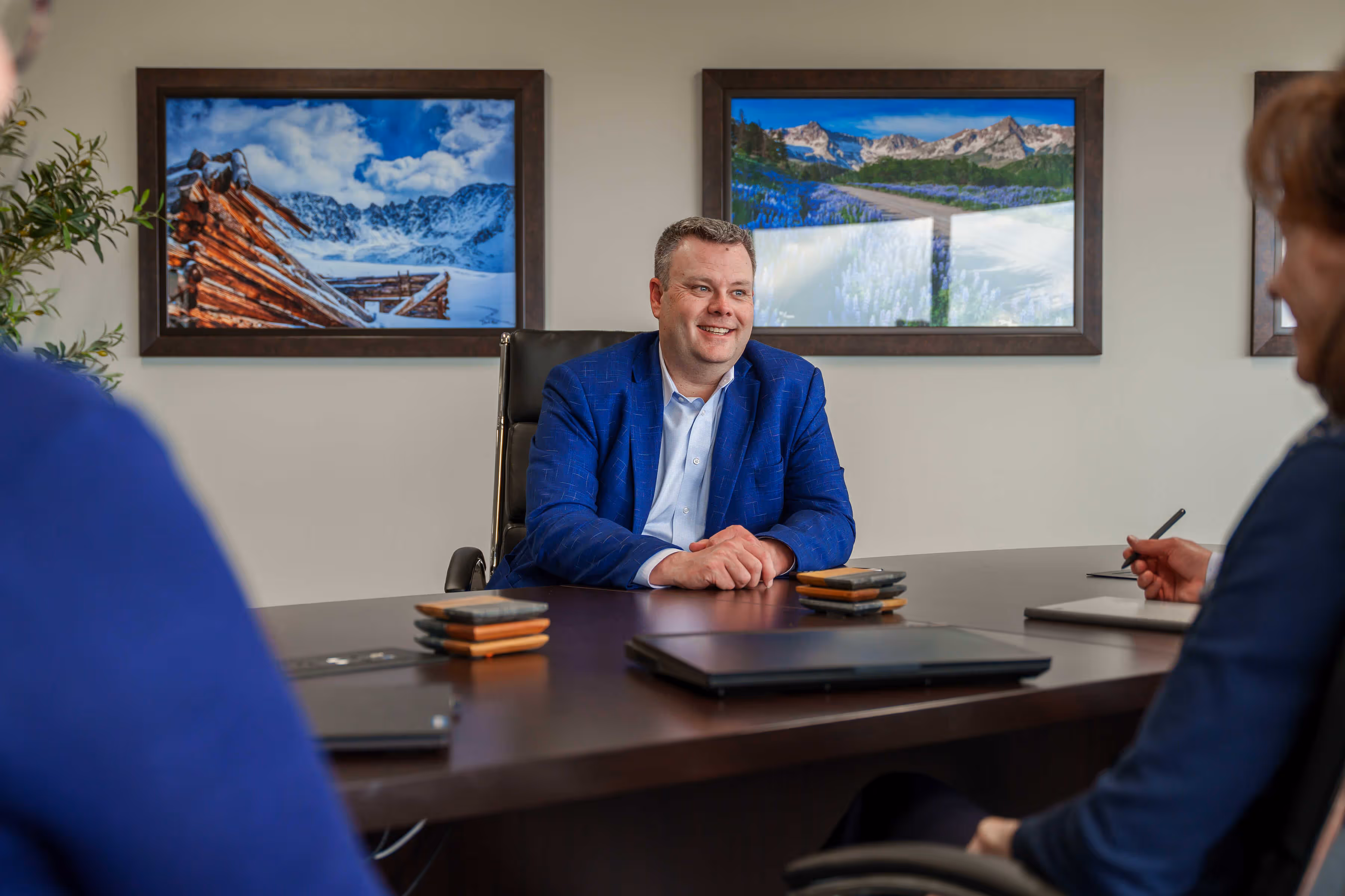 Man in blue blazer smiling and sitting at a table with two people in a meeting room with paintings.