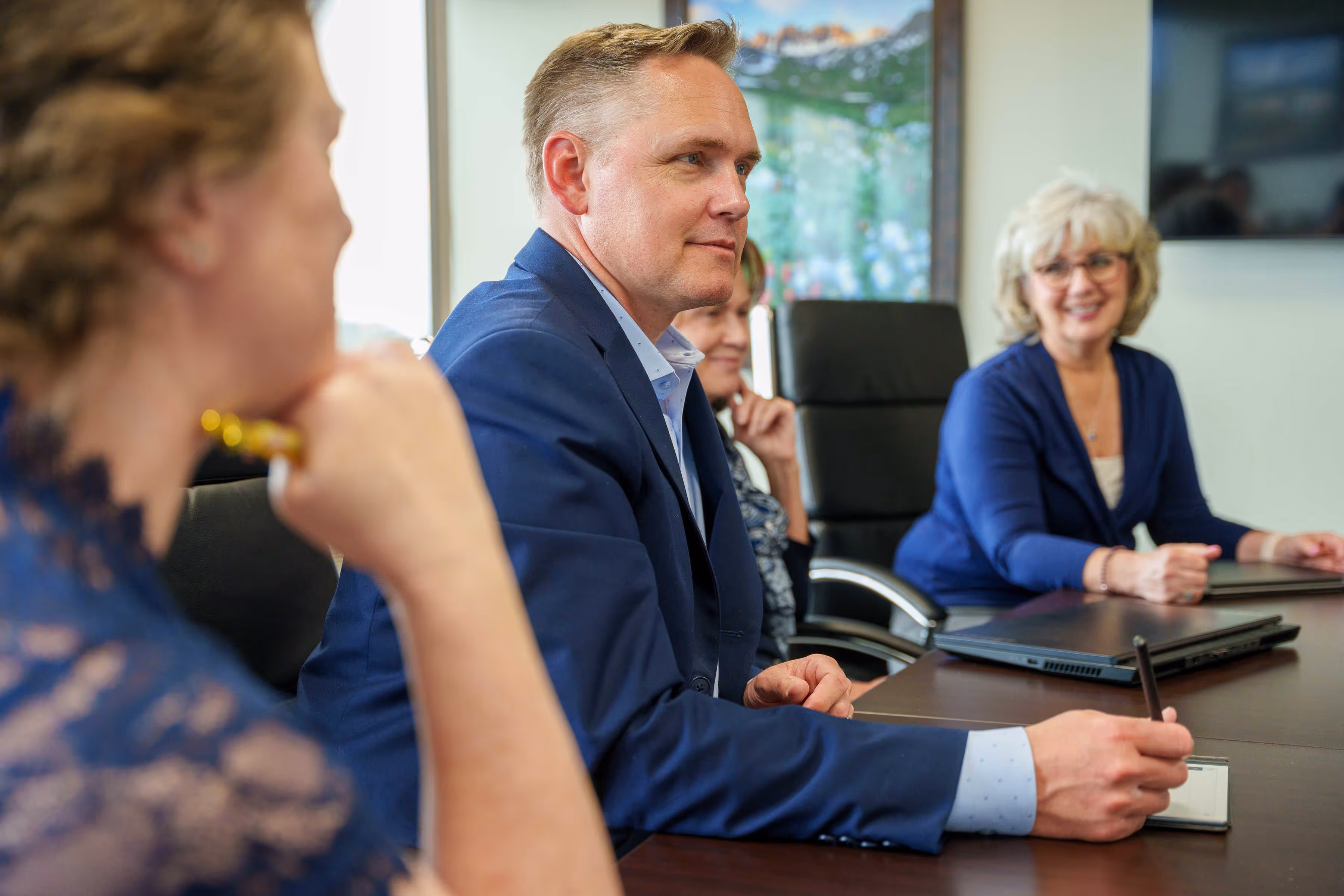 Four business professionals in a meeting room holding pens and talking around a conference table.