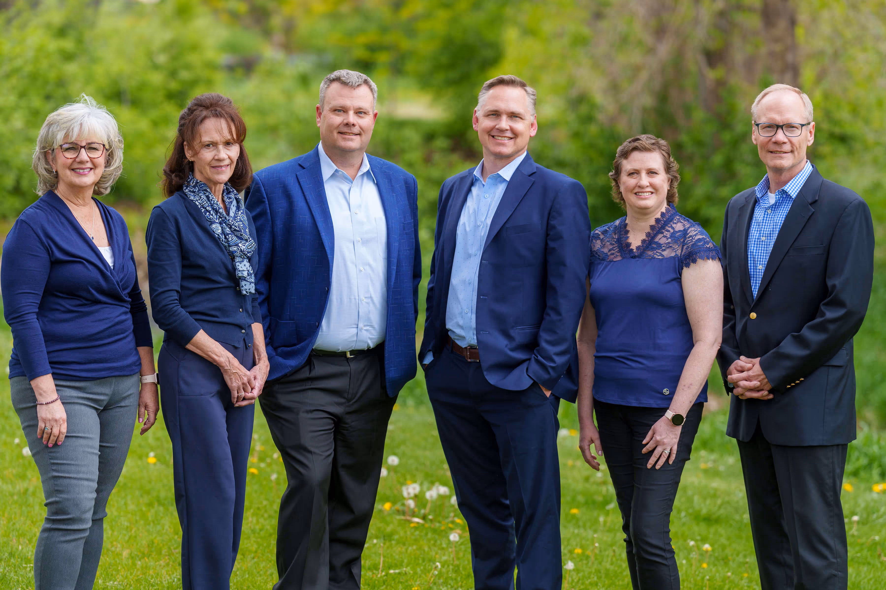 Group of six adults standing outdoors on grass with trees in the background, smiling at camera.