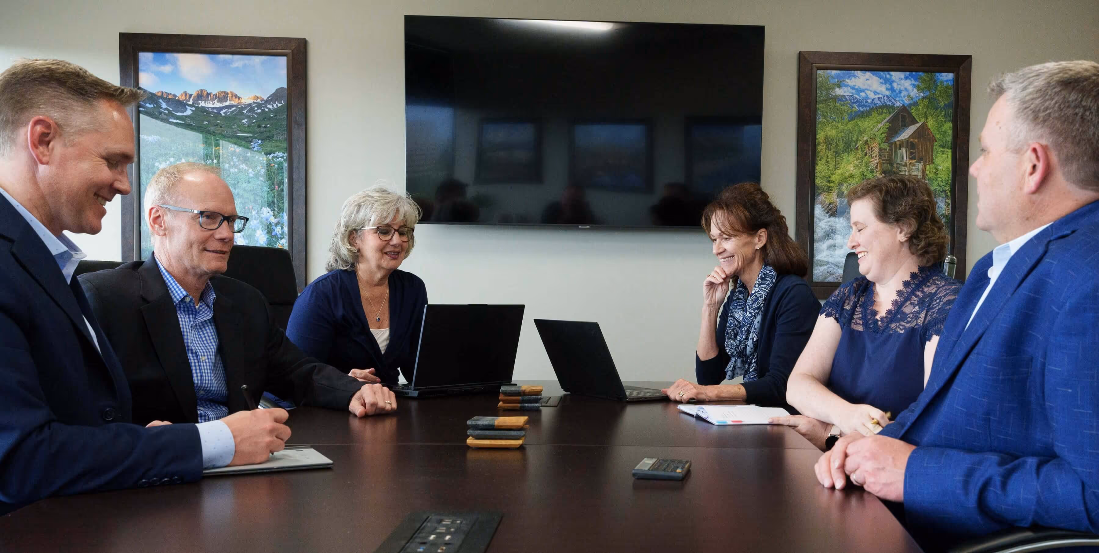 Six professionals in business attire sitting around a conference table with laptops and notepads.