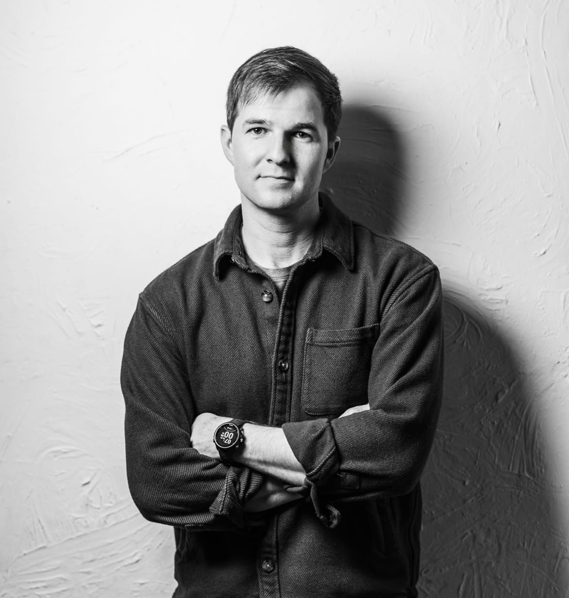 Black-and-white portrait of a man with short hair standing against a textured wall with arms crossed, wearing a buttoned shirt and a smartwatch.