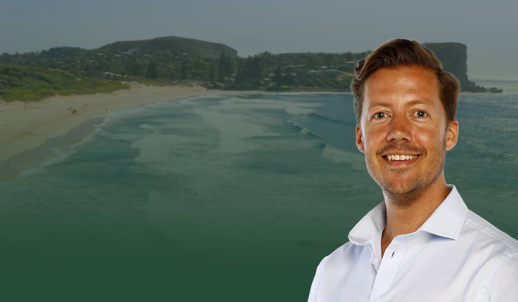 Smiling man in white shirt with a beach and forested hills in the background.