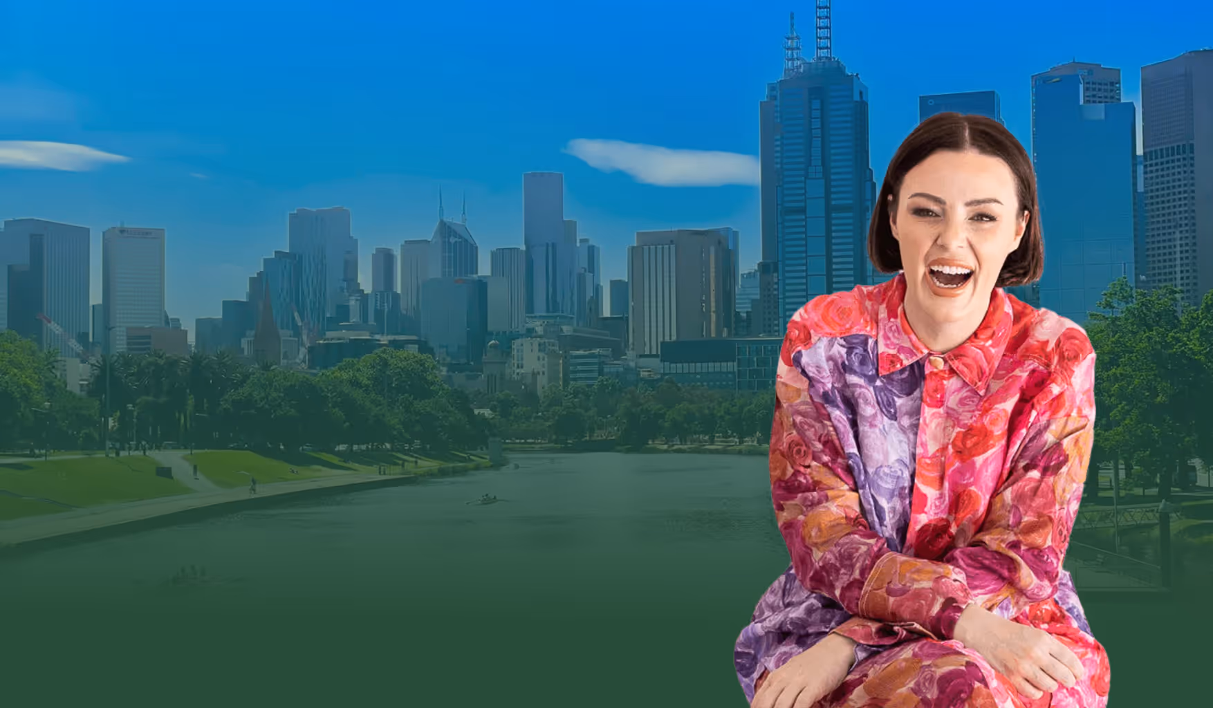 Smiling woman in colorful floral outfit sitting with city skyline and river in the background.