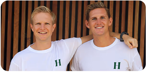 Two smiling men in white shirts with a green H logo, one with his arm around the other's shoulder.