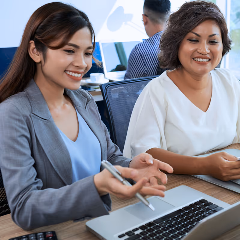 Two women smiling and discussing work while using a laptop in a modern office.