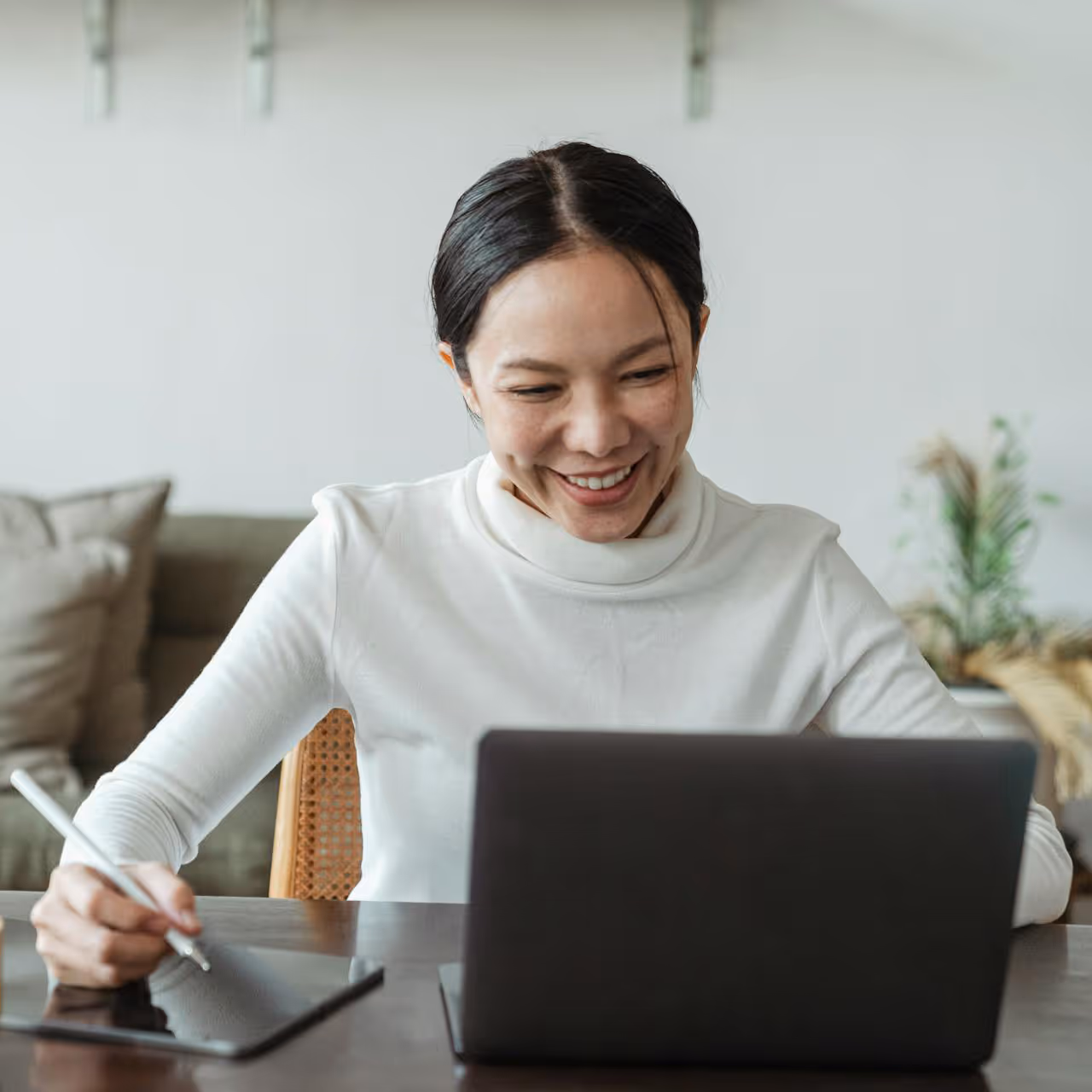 Smiling woman using a digital pen on a tablet while looking at her laptop screen indoors.