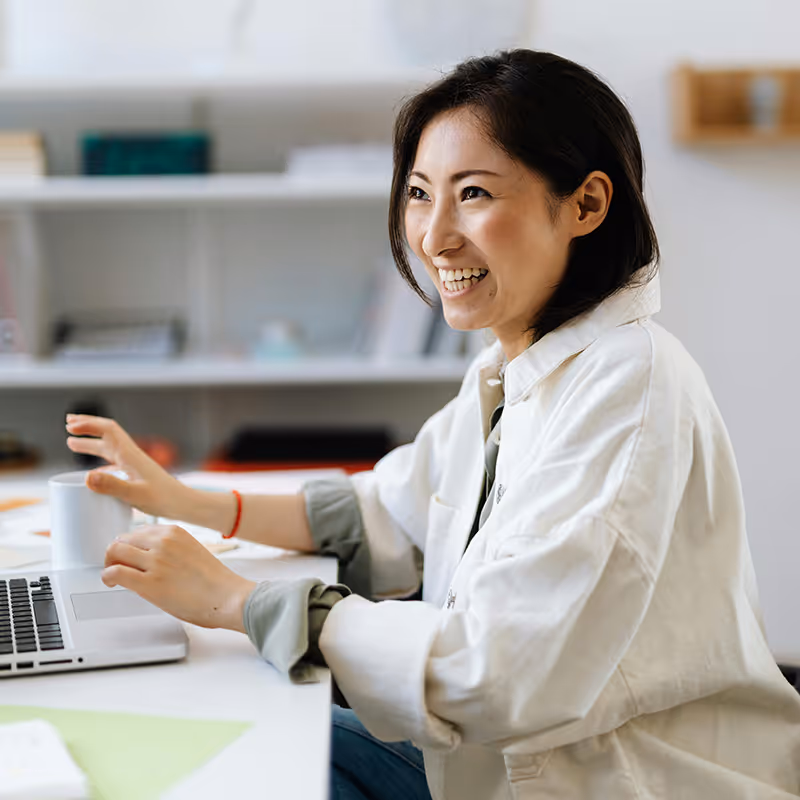 Smiling woman sitting at desk using laptop with coffee cup nearby in a bright room.