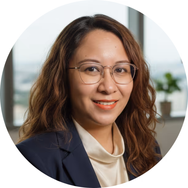 Smiling woman with wavy hair and glasses wearing a navy blazer and cream blouse in an office setting.
