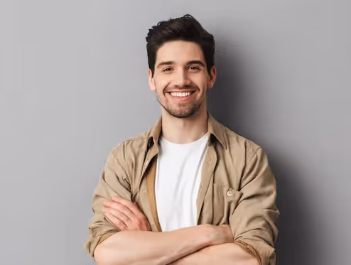 Nuori mies seisoo kädet ristissä ja hymyilee harmaata taustaa vasten.
