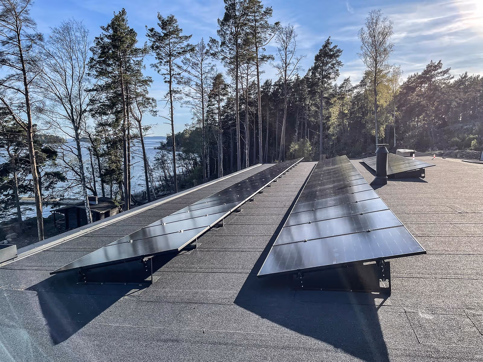 Kaksi aurinkopaneeliriviä litteällä katolla metsäisen merenrantamaiseman edessä kirkkaana päivänä.
