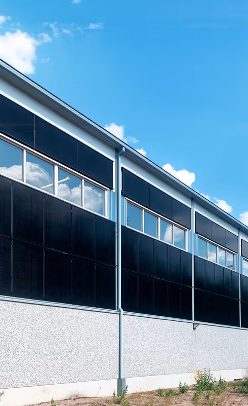 Teollinen rakennuksen seinä, jossa on mustia paneeleja ja ikkunoita, sininen taivas pilvineen taustalla.