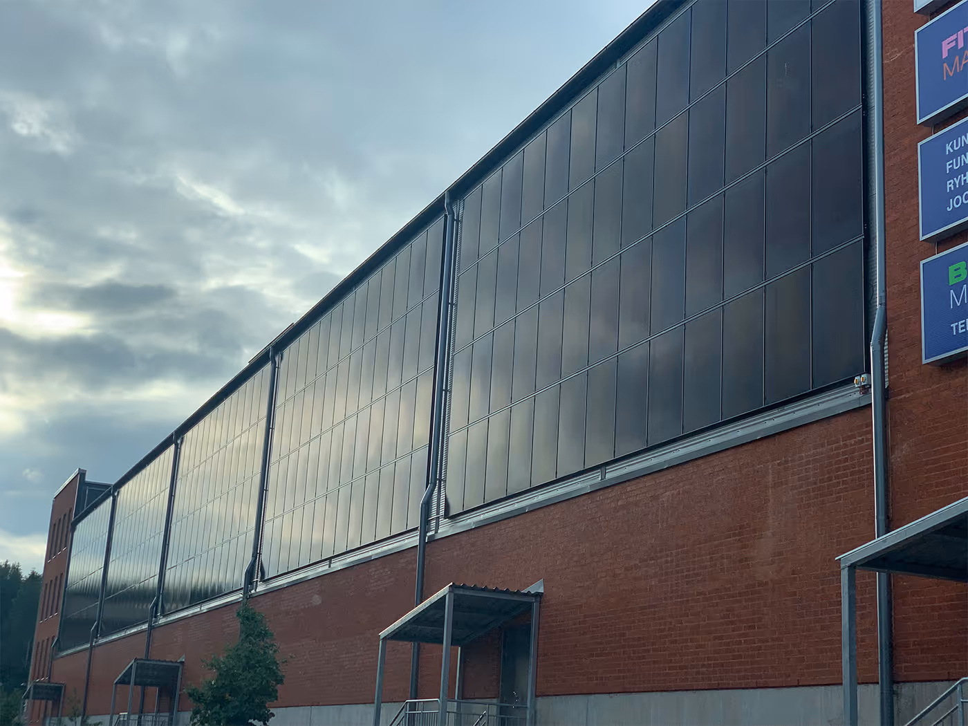 Suuri teollisuusrakennus, jonka yläosassa mustat aurinkopaneelit, alhaalla punatiiliseinää ja metallinen sisäänkäynti.