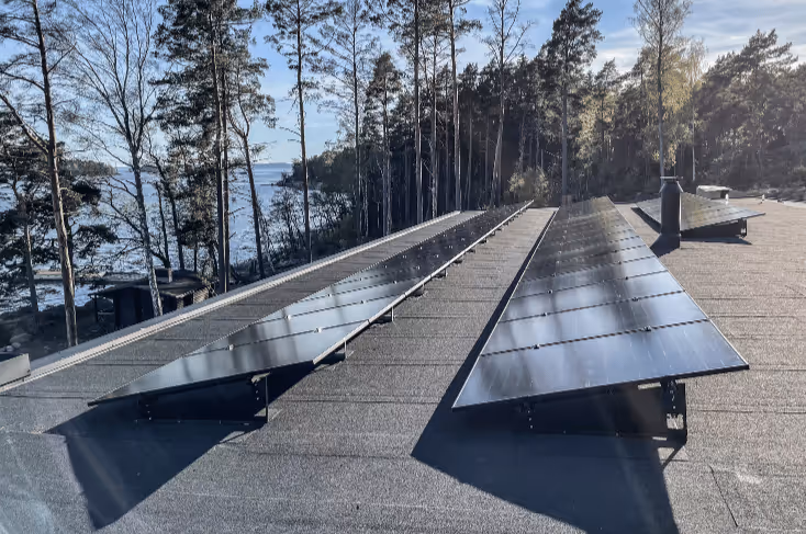 Kaksi rivillistä aurinkopaneeleja tasaisella katolla metsäisellä ja järvimaisemaisella taustalla.