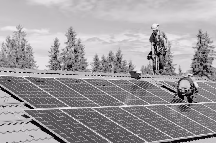 Kaksi työntekijää asentamassa aurinkopaneeleita talon katolle, taustalla mäntyjä ja pilvinen taivas.