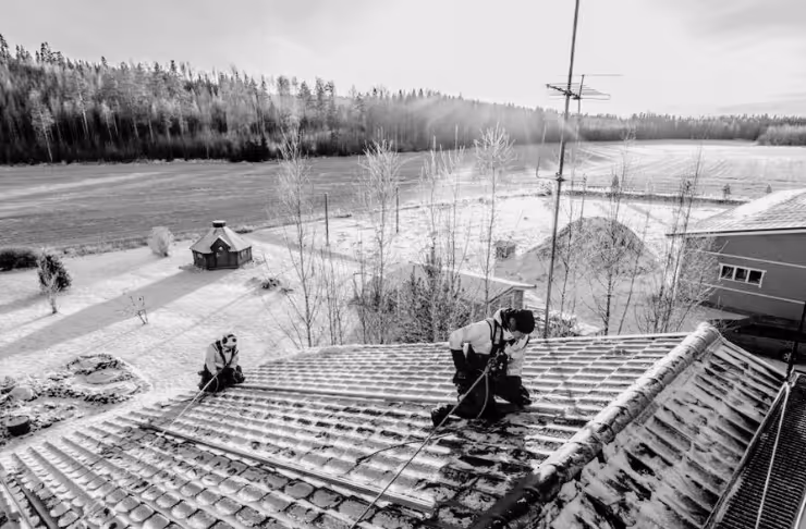 Kaksi henkilöä tekemässä Aurinpaneelienkattoasennusta lumisella kattotiilellä maalaismaisemassa.