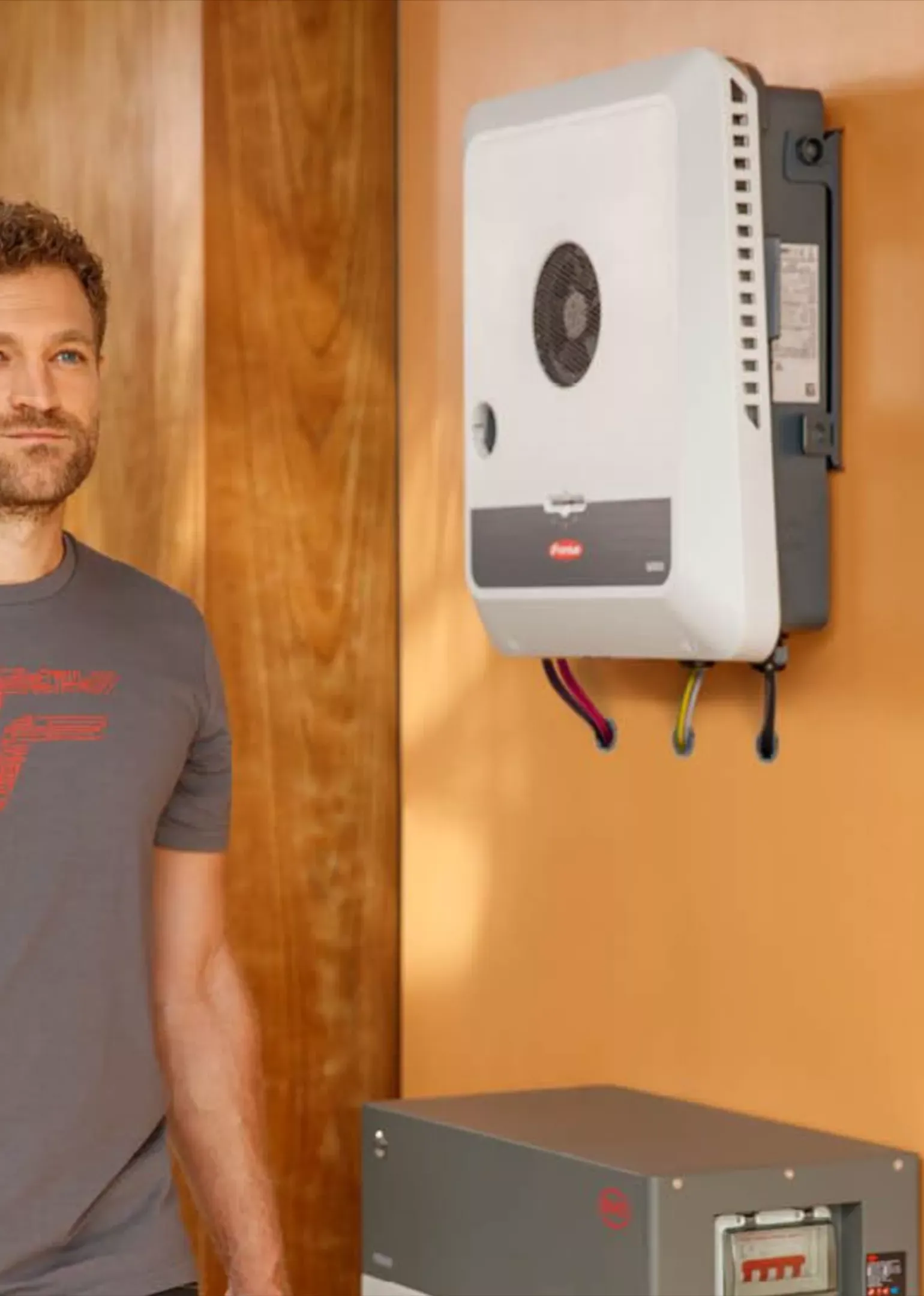 Mies harmaassa paidassa seisoo puuseinäisen huoneen vieressä, jossa on kiinnitetty valkoinen seinälaite ja harmaa sähkökeskus.