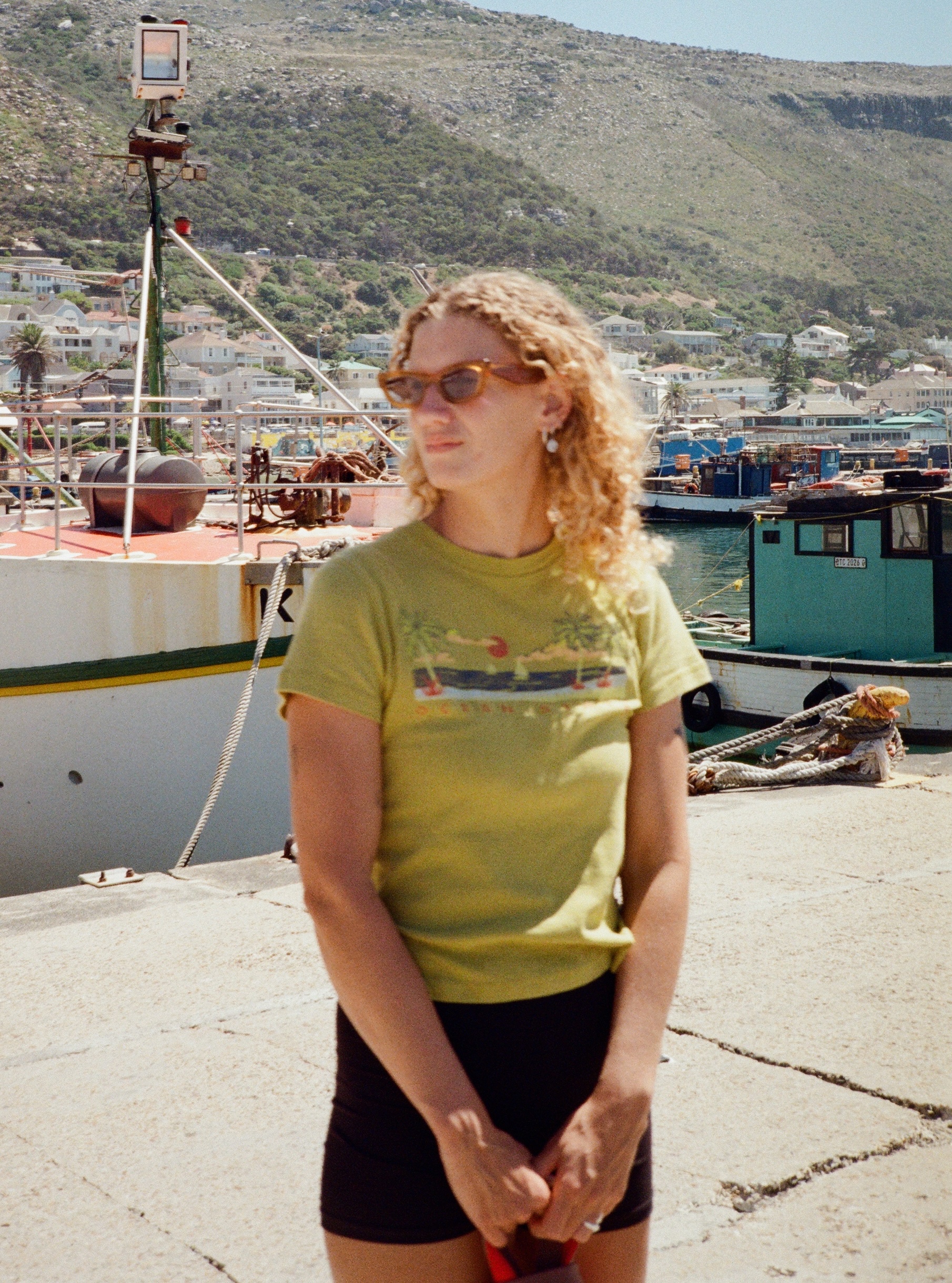 Shows Demi, A young woman in a geen top with orange sunglasses on. She has blond curly hair and is looking away from the camera. The background is a colourful harbour in Cape Town