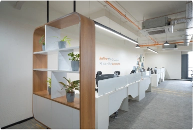 Modern Manubhai & Shah LLP office workspace with white desks, office chairs, and wooden shelf with potted plants.
