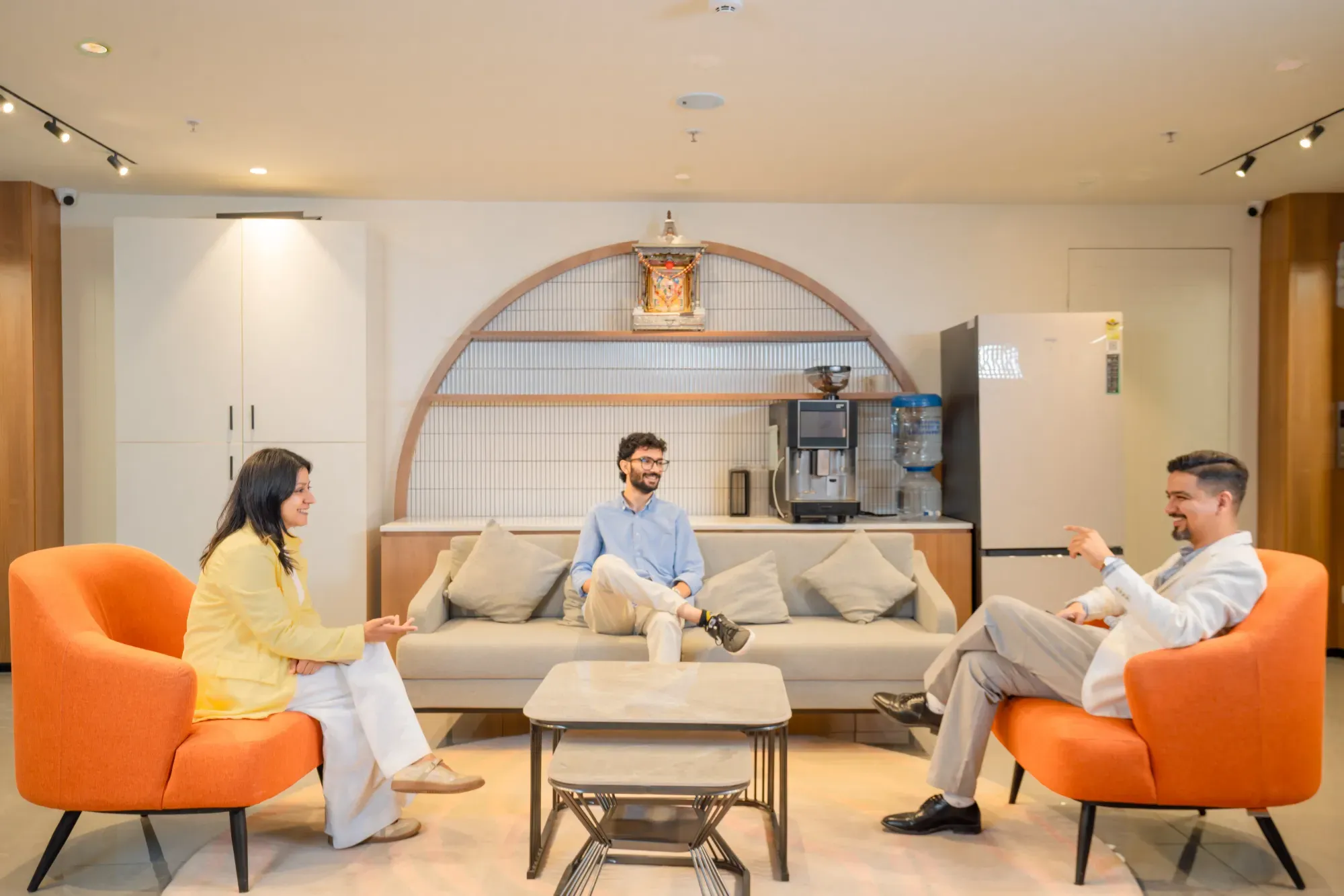 Two people sitting on a gray sofa talking, one person on orange chair, and one person walking by coffee area at Manubhai & Shah LLP office.