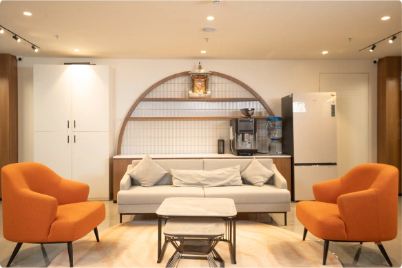 Modern living room with gray sofa, two orange armchairs, coffee table, and coffee machine on wooden shelf at Manubhai & Shah LLP office.