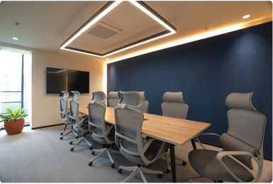 Modern conference room with a long wooden table, ergonomic chairs, a wall-mounted TV, and a potted plant at Manubhai & Shah LLP office.